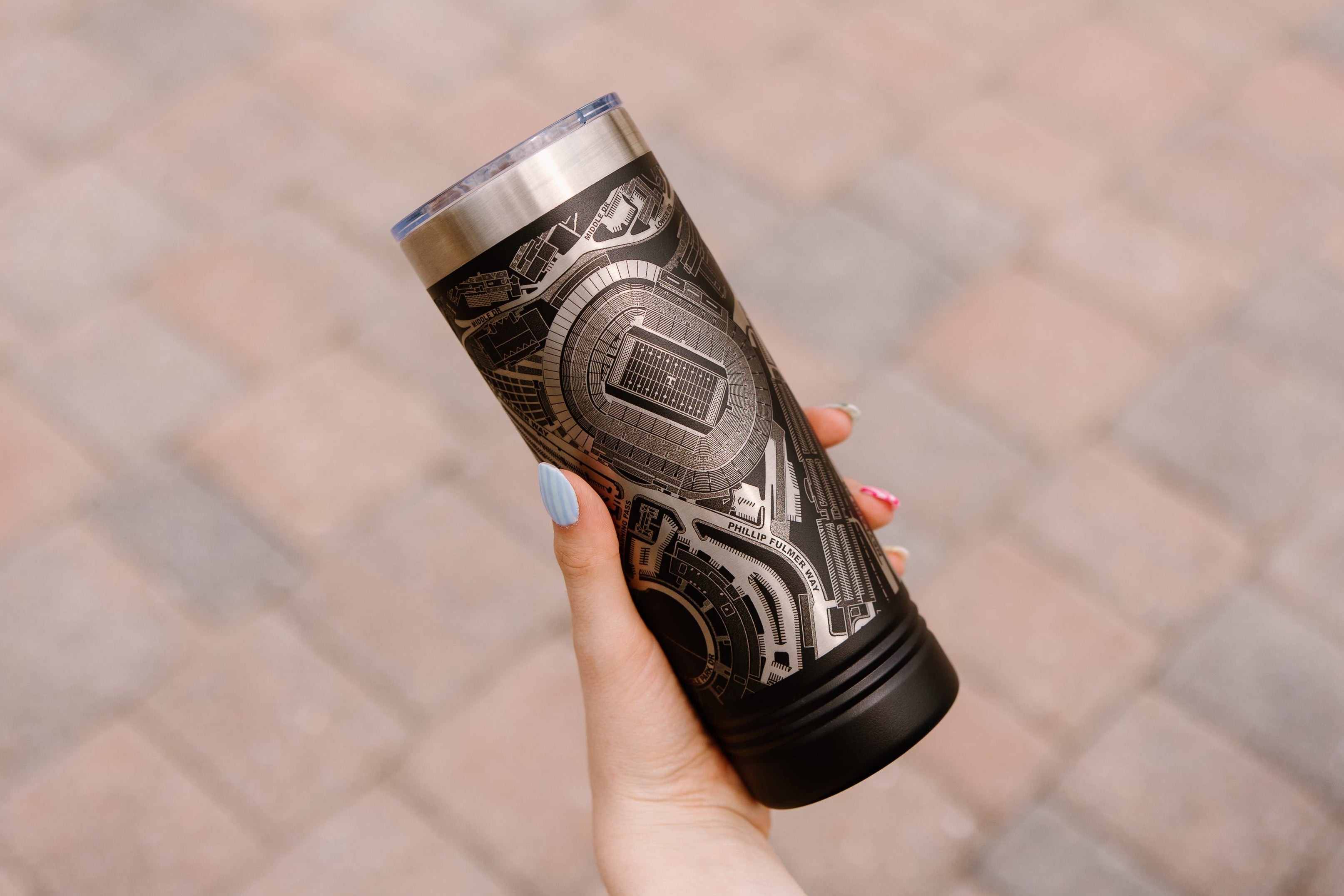 Person holding a silver and black tumbler with intricate design on a blurred pavement background