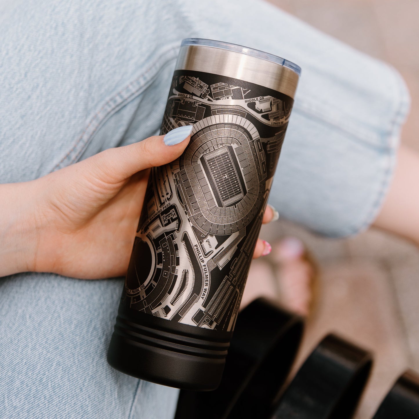 Person holding a tumbler with a detailed design, sitting on a bench.